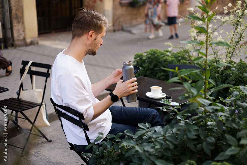 Sustainability city. Man drinks water from reusable metal bottle. Green city street cafe. Man wearing white shirt and using smartphone. Coffee cup on the table. Workation mobile internet on the go Sustainability city. Man drinks water from reusable metal bottle. Green city street cafe. Man wearing white shirt and using smartphone. Coffee cup on the table. Workation mobile internet on the go