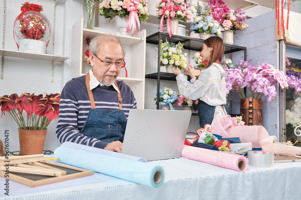 Asian old male florist owner working with a laptop on website for online business, a young beautiful female shopkeeper arranging fresh blossom behind, colorful flower shop store, e-commerce SME. Asian old male florist owner working with a laptop on website for online business, a young beautiful female shopkeeper arranging fresh blossom behind, colorful flower shop store, e-commerce SME.