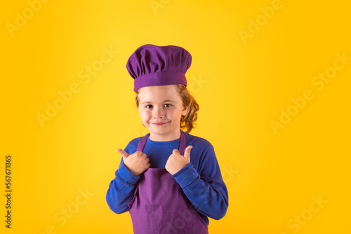 Child cooking, little chef prepares food. Kid boy in chefs hat and apron on yellow studio isolated background.