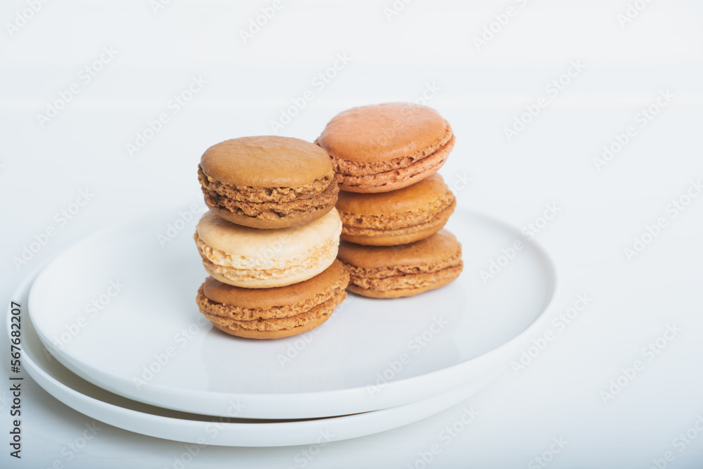 Stack of delicious freshly baked caramel, coffee and vanilla flavoured macaron confections on white plate against white background. Copy space. French sweet food speciality.