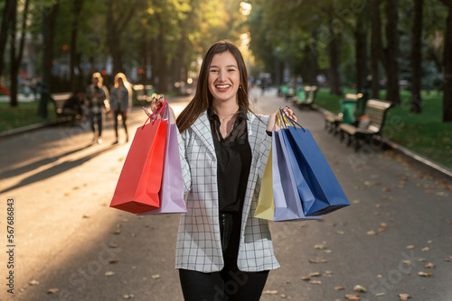 Fashionable beautiful young caucasian woman carrying colorful shopping bags on alley of the park background.
