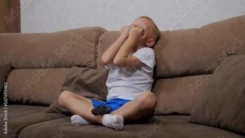 Close-up of a sad little blond boy sitting on the sofa at home, holding a remote control. The child is punished and does not watch TV. photo