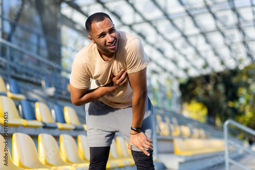 Sick athlete after jogging and vigorous exercise has severe chest pain, heart attack in young afro american athlete, man holding hands on chest in stadium sunny afternoon.