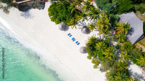 Aerial view of umbrellas, palms on the sandy beach of Indian Ocean at sunny day. Summer holiday in Zanzibar, Africa. Tropical landscape with palm trees, parasols, boats, yachts, blue water. Top view