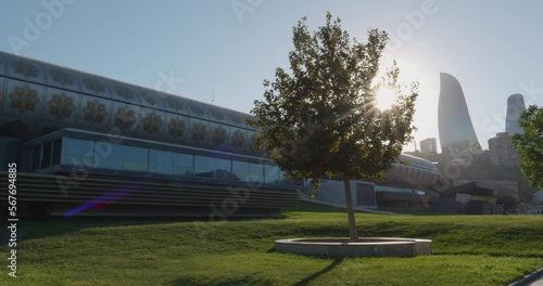 Azerbaijan, Baku. Building of the carpet museum and the tower of Flame Towers complex against the backdrop of a blue sky with a bright disk of the shining sun through dense branches of a green tree