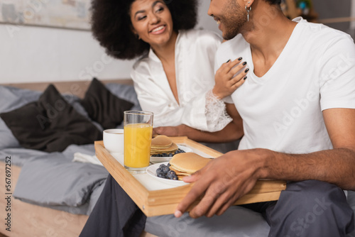 smiling african american woman touching boyfriend holding tray with delicious pancakes and orange juice in bedroom