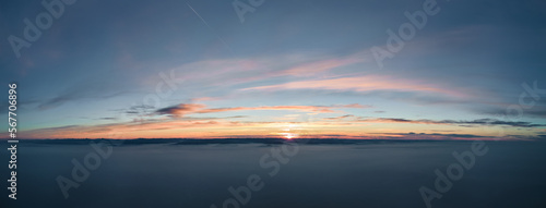 Aerial view of colorful sunset over white dense foggy clouds cover with distant dark silhouettes of mountain hills on horizon