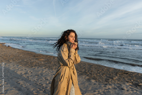 Photography cheerful woman in stylish beige trench coat walking on beach in Barcelona