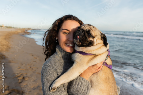 Canvas Print cheerful and curly young woman holding pug dog on beach near sea in Barcelona