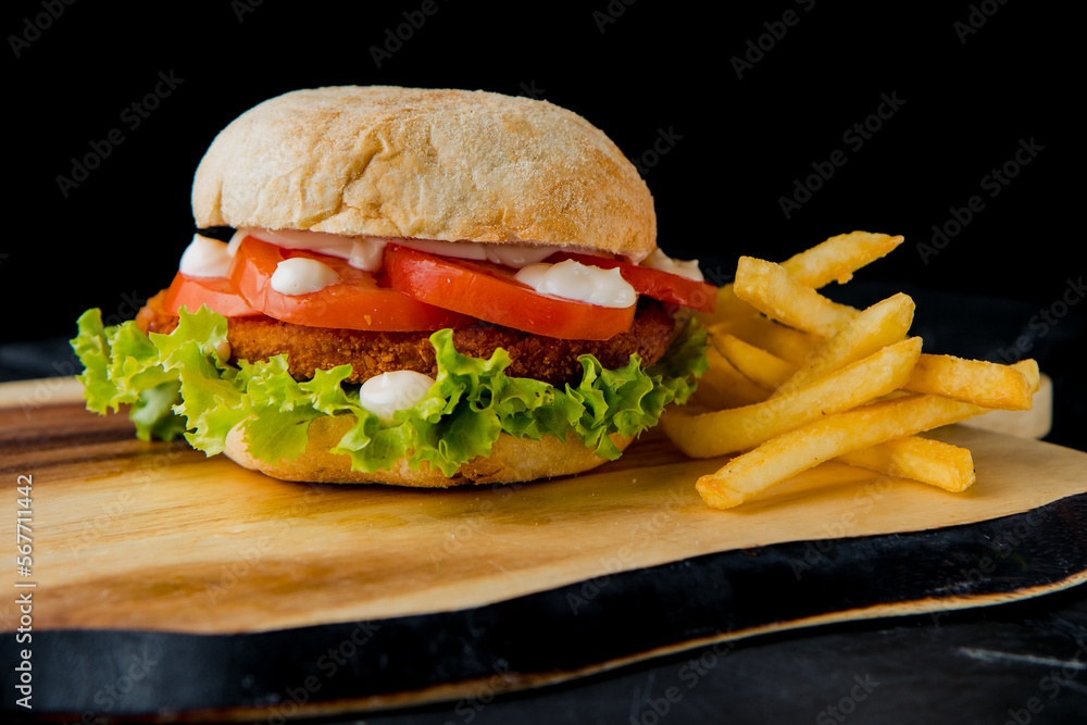 Classic hamburger with pork cutlet, tomato, salad leaves, white sauce with french fries on the wooden plate on the dark background Classic hamburger with pork cutlet, tomato, salad leaves, white sauce with french fries on the wooden plate on the dark background