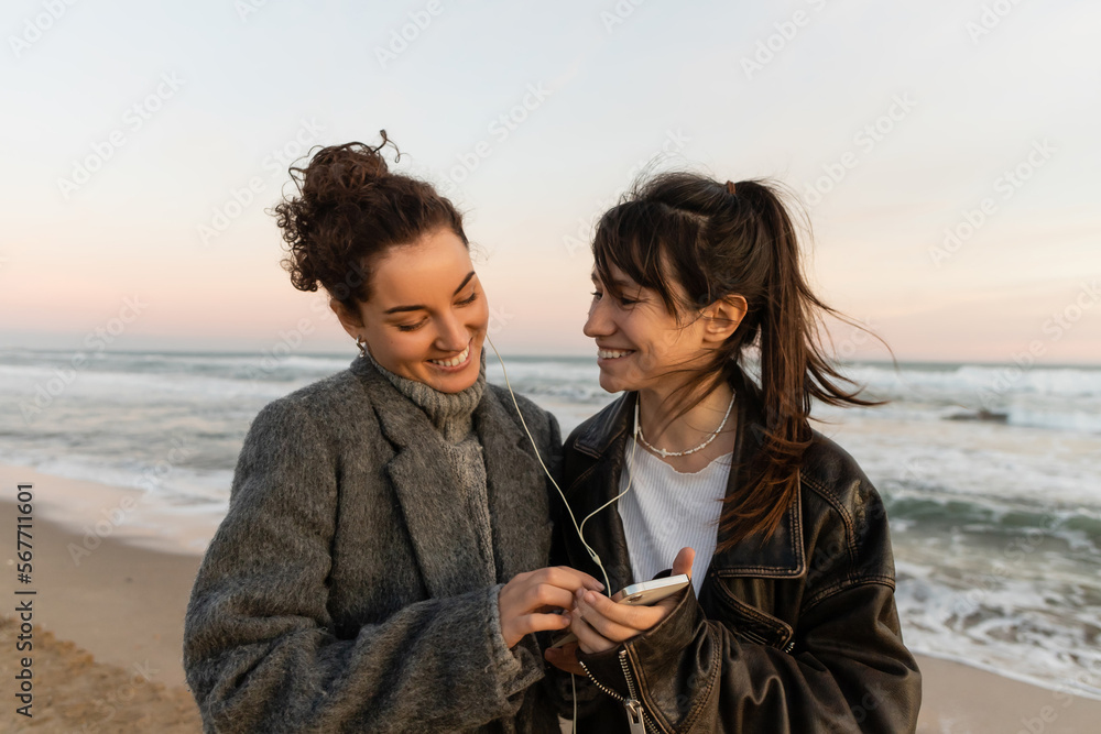 Smiling friends listening music in wired earphones and holding smartphone on beach.