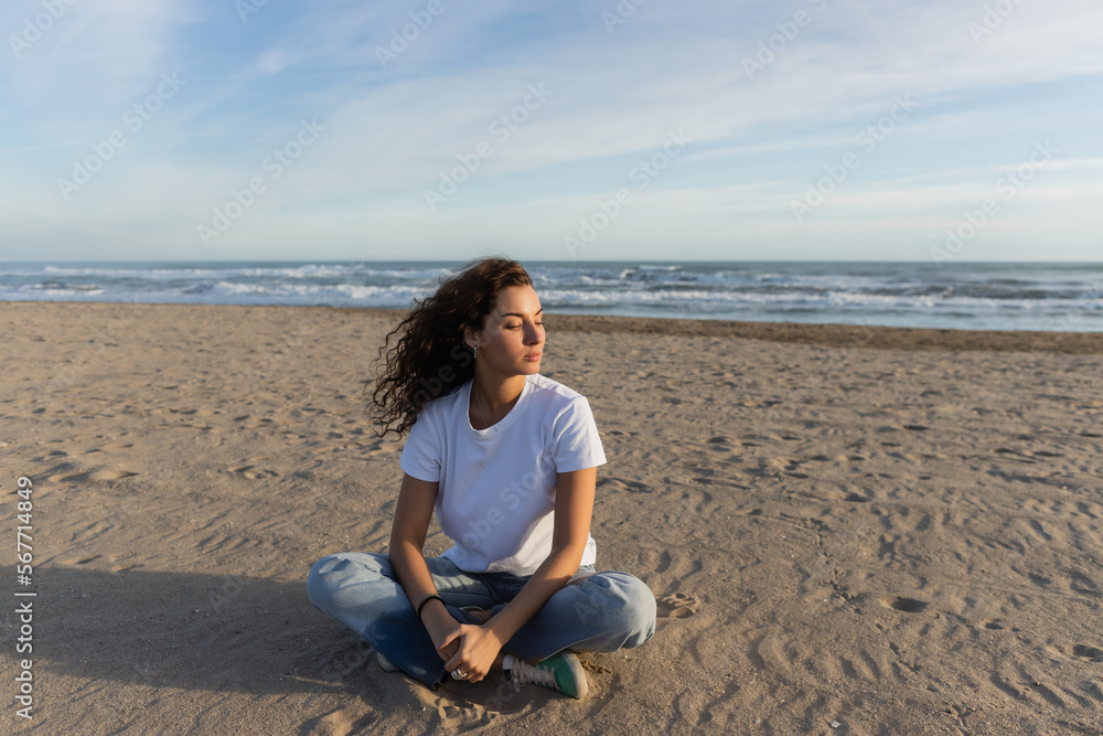curly brunette woman sitting in blue jeans and white t-shirt sitting on sandy beach in Spain.