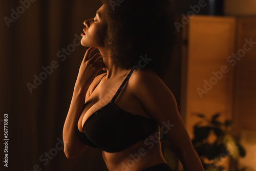 side view of young african american woman with sexy bust touching neck while posing at night in bedroom