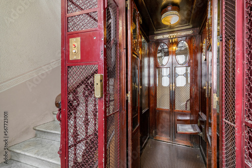 Empty retro style elevator with red metal bars, doors on both sides, inside with dark polished wood walls. Elevator is illuminated by a lamp with a yellow shade that adds mystery and atmosphere.