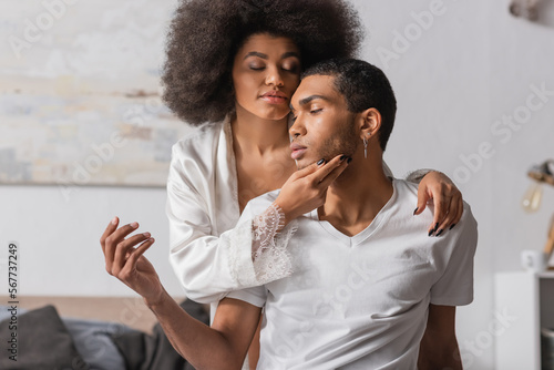 sexy african american woman with curly hair and closed eyes touching face of young boyfriend in bedroom