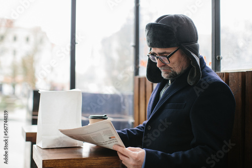 A gray-haired man in warm clothes reads a fresh newspaper and drinks hot tea or coffee on the street.