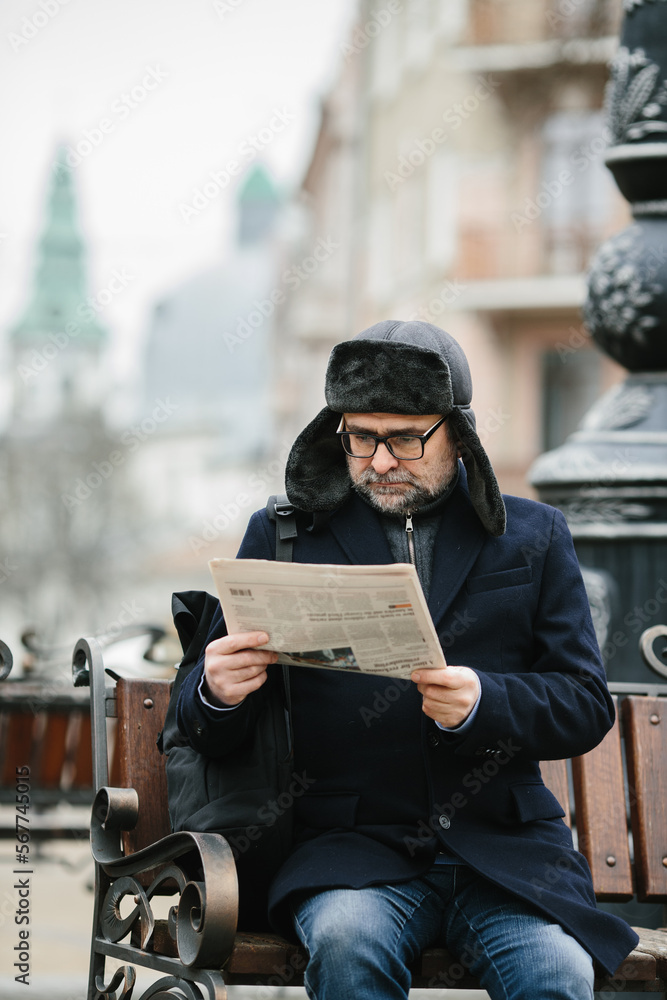 An elderly man enjoys a cold winter morning in the city. A man sits on a bench in the square, reads a newspaper and drinks coffee.