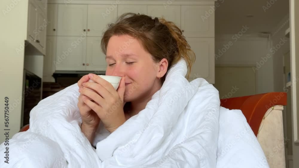 Young happy beautiful sleepy lazy woman is drinking from cup tea or coffee in early morning covering herself in blanket in bed in bedroom, smiling Young happy beautiful sleepy lazy woman is drinking from cup tea or coffee in early morning covering herself in blanket in bed in bedroom, smiling