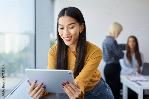 Smiling young woman at office work using digital tablet with coworkers in the background