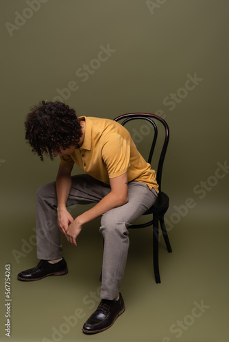full length of african american man in yellow polo shirt and black leather shoes sitting on chair with bowed head on grey green background © LIGHTFIELD STUDIOS