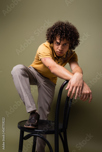 tattooed african american man in yellow polo shirt posing with chair while looking at camera on grey green background © LIGHTFIELD STUDIOS