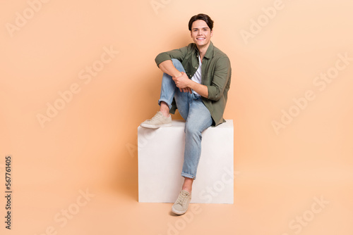 Full length photo cadre of young positive brunet hair man sitting comfortable cube platform wear casual outfit isolated on beige color background