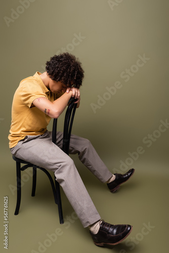 full length of tattooed african american man in yellow polo shirt and pants sitting on chair with bowed head on grey green background © LIGHTFIELD STUDIOS