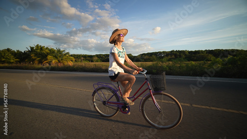 Wide angle side view of pretty mature senior woman biking into the sun wearing sunglasses hat ethnic attire in a tropical setting with palm trees.