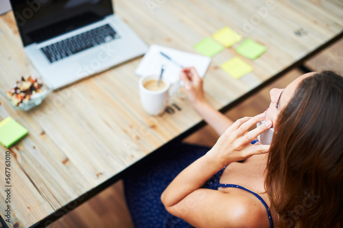 woman during phone call. Overhead of brunette woman in dress sitting at desk with stick notes  notebook  laptop and mug of coffee talking on cell phone.