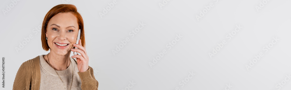 Cheerful red haired woman talking on cellphone isolated on grey, banner.
