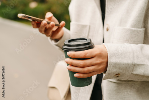 Close up shot of female hands holding phone and a cup of hot to go coffee outdoors.