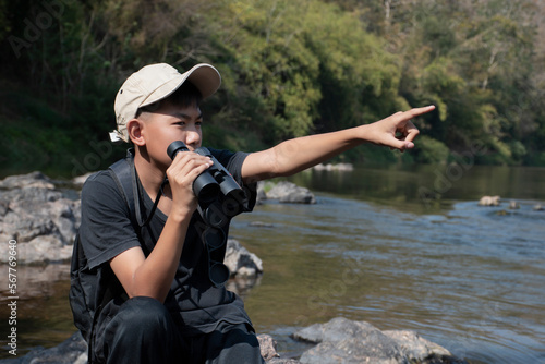 Asian boy wearing black t-shirt holding a binoculars sitting on stone by the river, pointing to birds on trees in national park to observe fish in the river and birds on tree branches and on sky.