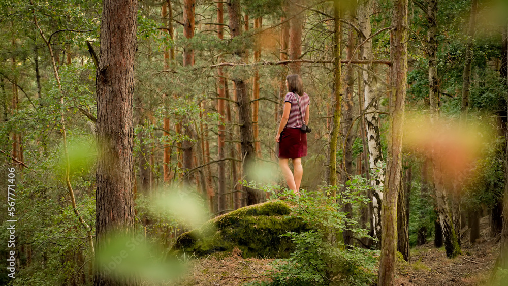 A young woman in a red skirt is stand on a rock in a forest. In the bokeh foreground are some blured leaves.
