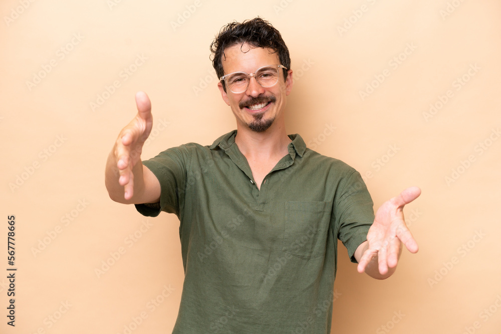 Young caucasian man isolated on beige background presenting and inviting to come with hand Young caucasian man isolated on beige background presenting and inviting to come with hand