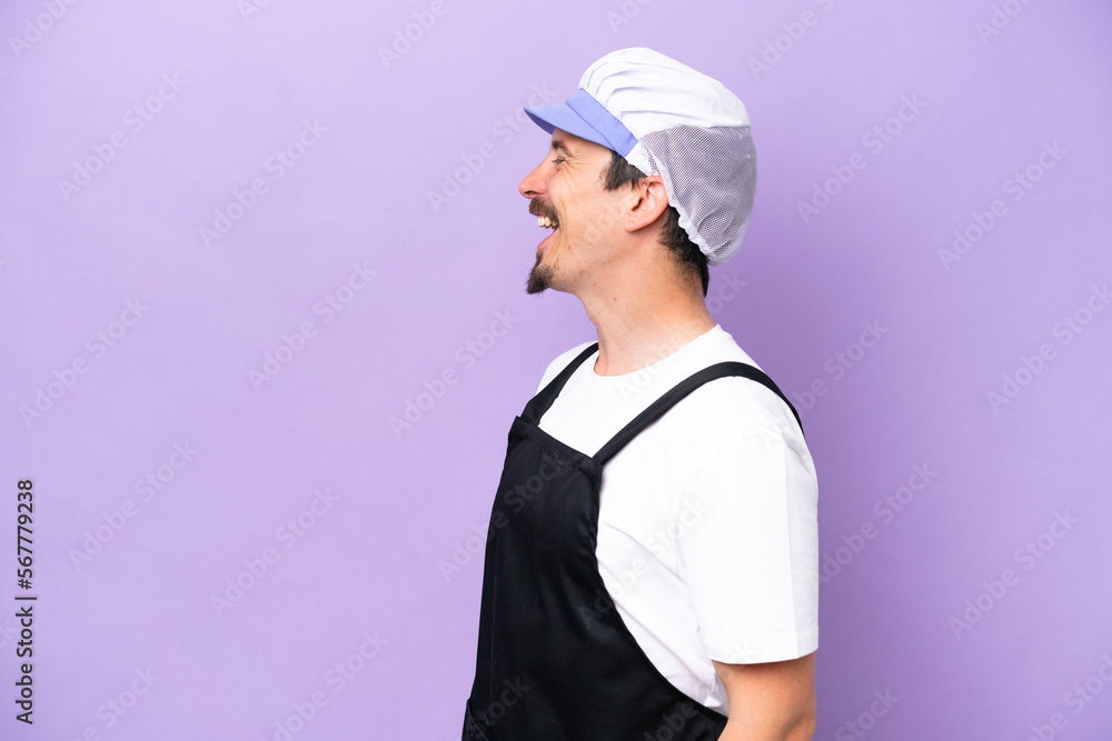 Fishmonger man wearing an apron isolated on purple background laughing in lateral position Fishmonger man wearing an apron isolated on purple background laughing in lateral position
