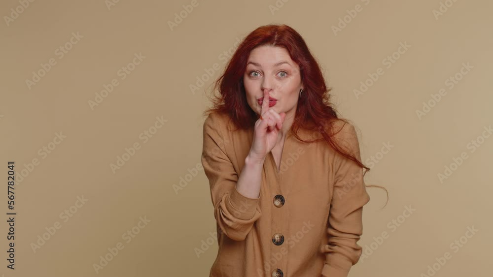 Shh be quiet please. Portrait of millennial woman 20 years old presses index finger to lips makes silence gesture sign do not tells secret. Young lovely redhead girl posing on beige studio background Shh be quiet please. Portrait of millennial woman 20 years old presses index finger to lips makes silence gesture sign do not tells secret. Young lovely redhead girl posing on beige studio background
