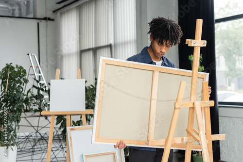 Young african american artist holding canvas near wooden easel in studio.