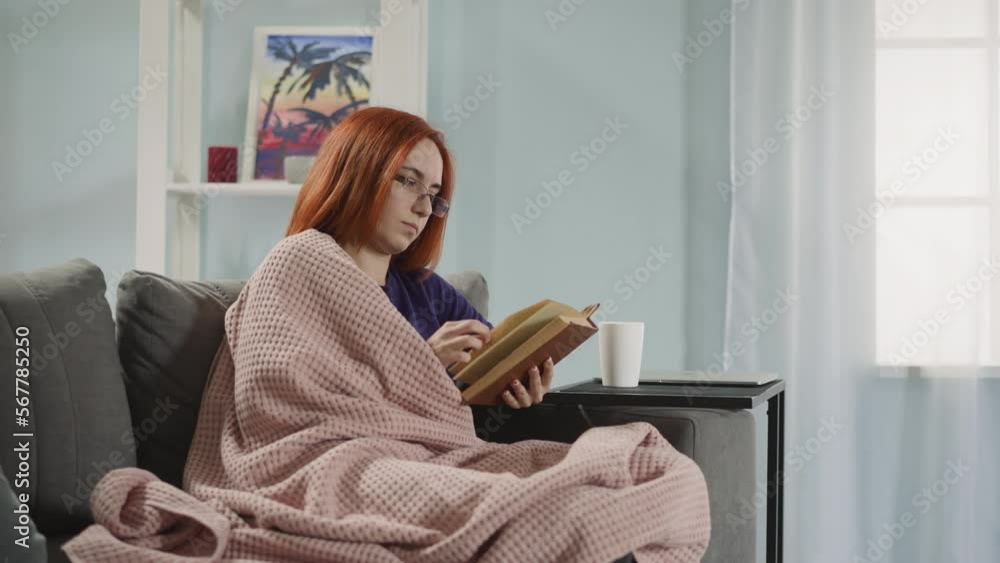 Red-haired woman in glasses reads interesting love story sitting on couch near bright window. Focused lady covered with plaid flips pages of book Red-haired woman in glasses reads interesting love story sitting on couch near bright window. Focused lady covered with plaid flips pages of book