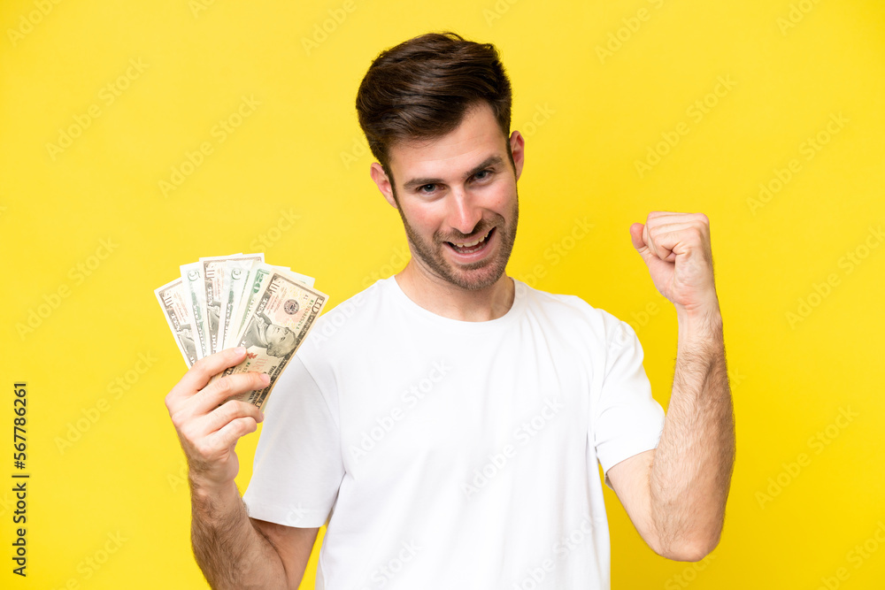Young caucasian man taking a lot of money isolated on yellow background celebrating a victory Young caucasian man taking a lot of money isolated on yellow background celebrating a victory