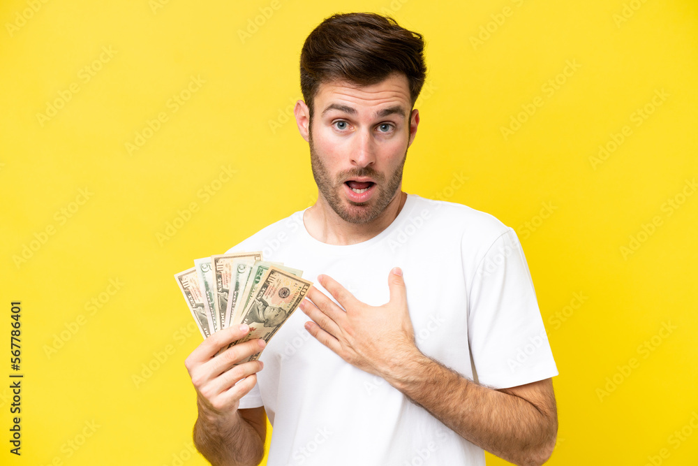 Young caucasian man taking a lot of money isolated on yellow background surprised and shocked while looking right Young caucasian man taking a lot of money isolated on yellow background surprised and shocked while looking right