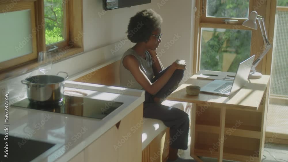 Medium long shot of young black woman sitting in kitchen at home and chatting via online video call on laptop Medium long shot of young black woman sitting in kitchen at home and chatting via online video call on laptop