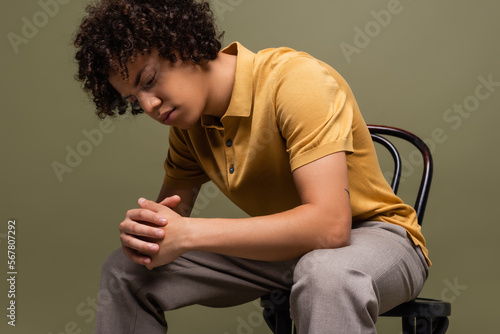 young african american man with curly hair wearing yellow polo shirt and sitting on chair with clenched hands isolated on grey © LIGHTFIELD STUDIOS
