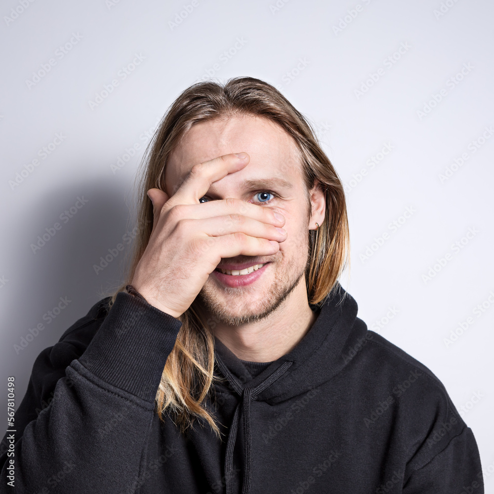 Portrait of a real smiling young blond man with blue eyes and long hair on a plain background, looking through his fingers straight into the camera, his face is covered with his hand Portrait of a real smiling young blond man with blue eyes and long hair on a plain background, looking through his fingers straight into the camera, his face is covered with his hand