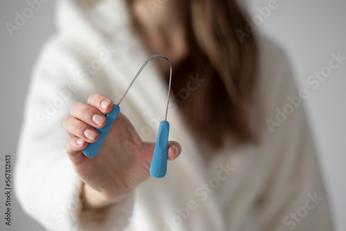 Metal tongue scraper tool in hand against the background of a woman in a bathrobe.