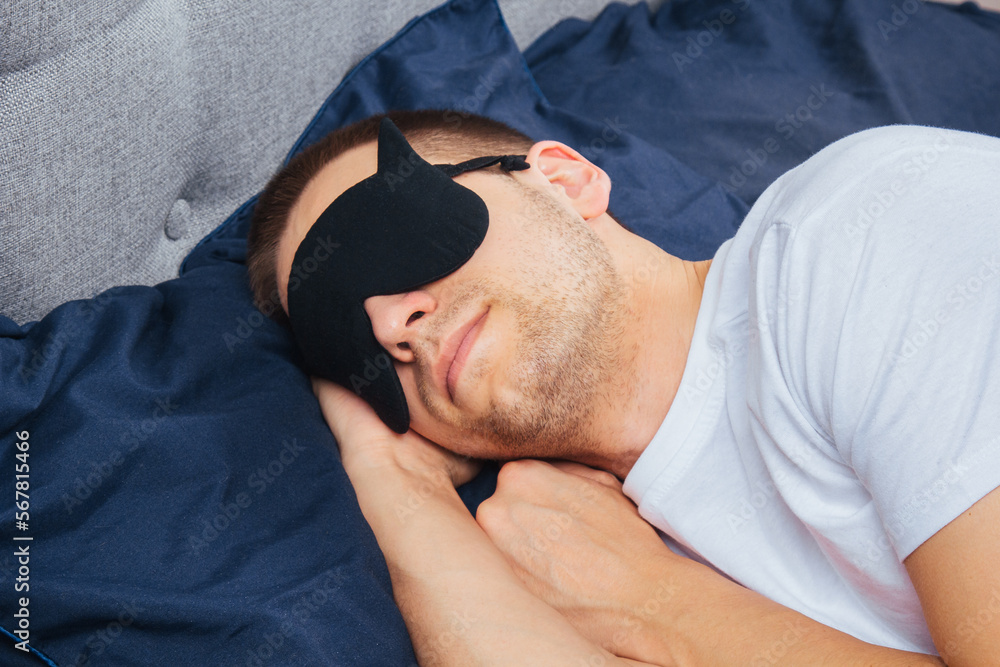 Side view of a young smiling cheerful man in a sleep mask in pajamas resting, relaxing at home, wrapping in a blanket, walking isolated on a dark blue background. Side view of a young smiling cheerful man in a sleep mask in pajamas resting, relaxing at home, wrapping in a blanket, walking isolated on a dark blue background.