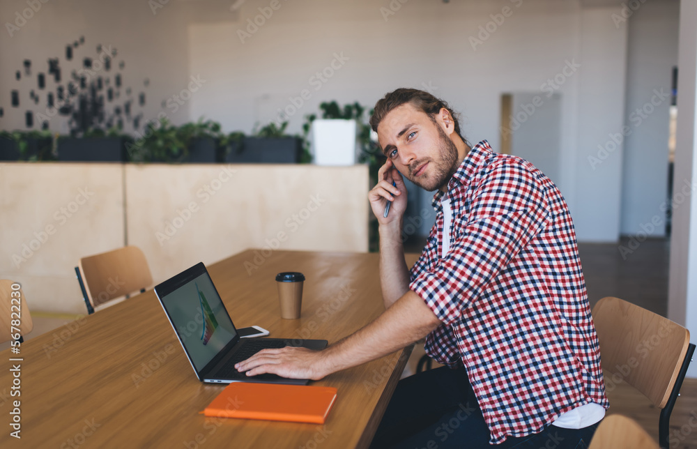 Portrait of skilled Caucaisan IT professional looking at camera during distance job in coworking space, millennial man in smart casual wear posing at table desktop with modern laptop computer Portrait of skilled Caucaisan IT professional looking at camera during distance job in coworking space, millennial man in smart casual wear posing at table desktop with modern laptop computer