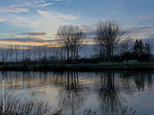 River Scheldt with bare winter trees reflecting in the water under a colorful cloudy sky in Merelbeke, Flandeers, Belgium