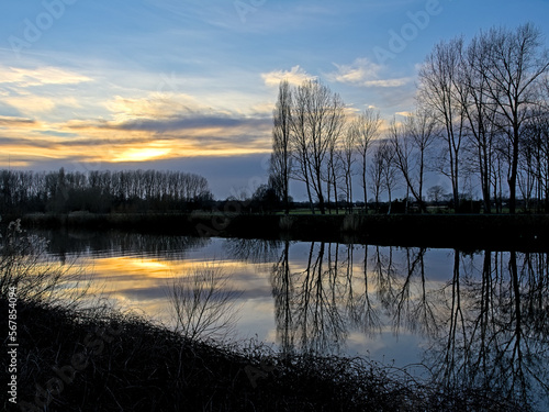 River Scheldt with bare winter trees reflecting in the water under a colorful cloudy sky in Merelbeke, Flandeers, Belgium