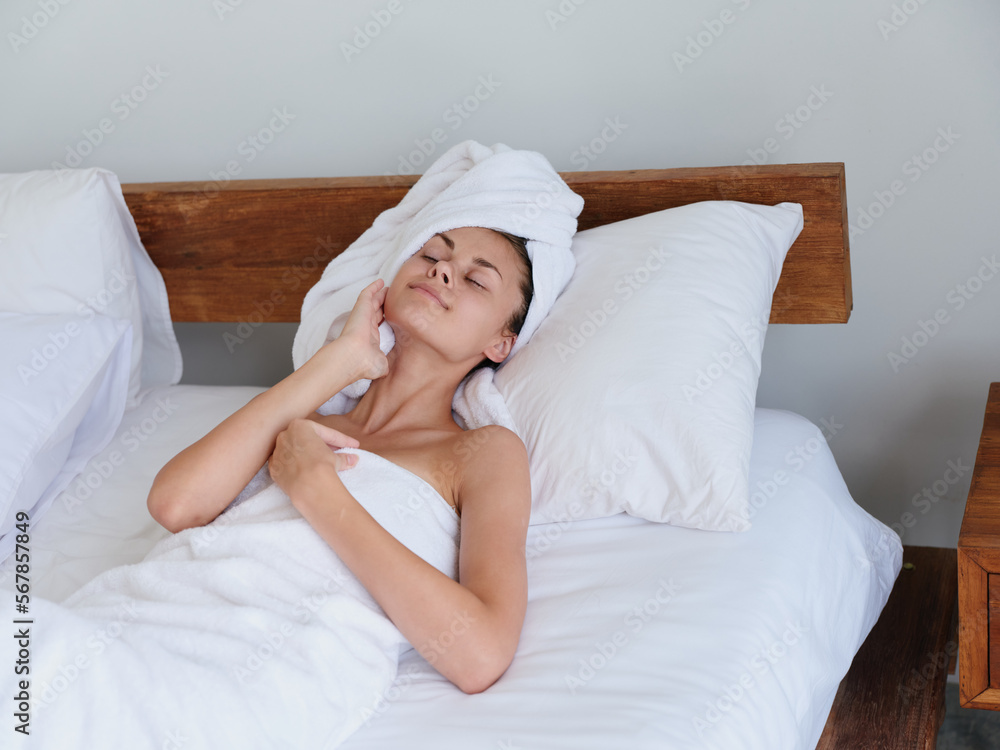 A woman lies on the bed after a shower with wet hair with a towel on her head in the bedroom and strokes her body skin, a cute smile with teeth and weekend rest in bed, women's beauty day, lifestyle A woman lies on the bed after a shower with wet hair with a towel on her head in the bedroom and strokes her body skin, a cute smile with teeth and weekend rest in bed, women's beauty day, lifestyle
