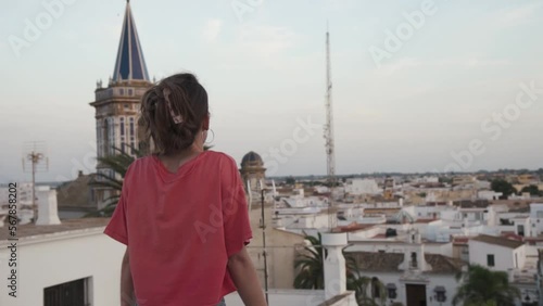 Young attractive girl contemplates a church and the andalalusian city from a balcony in Chipiona, Cadiz, Spain. She wears shorts and a pink shirt.